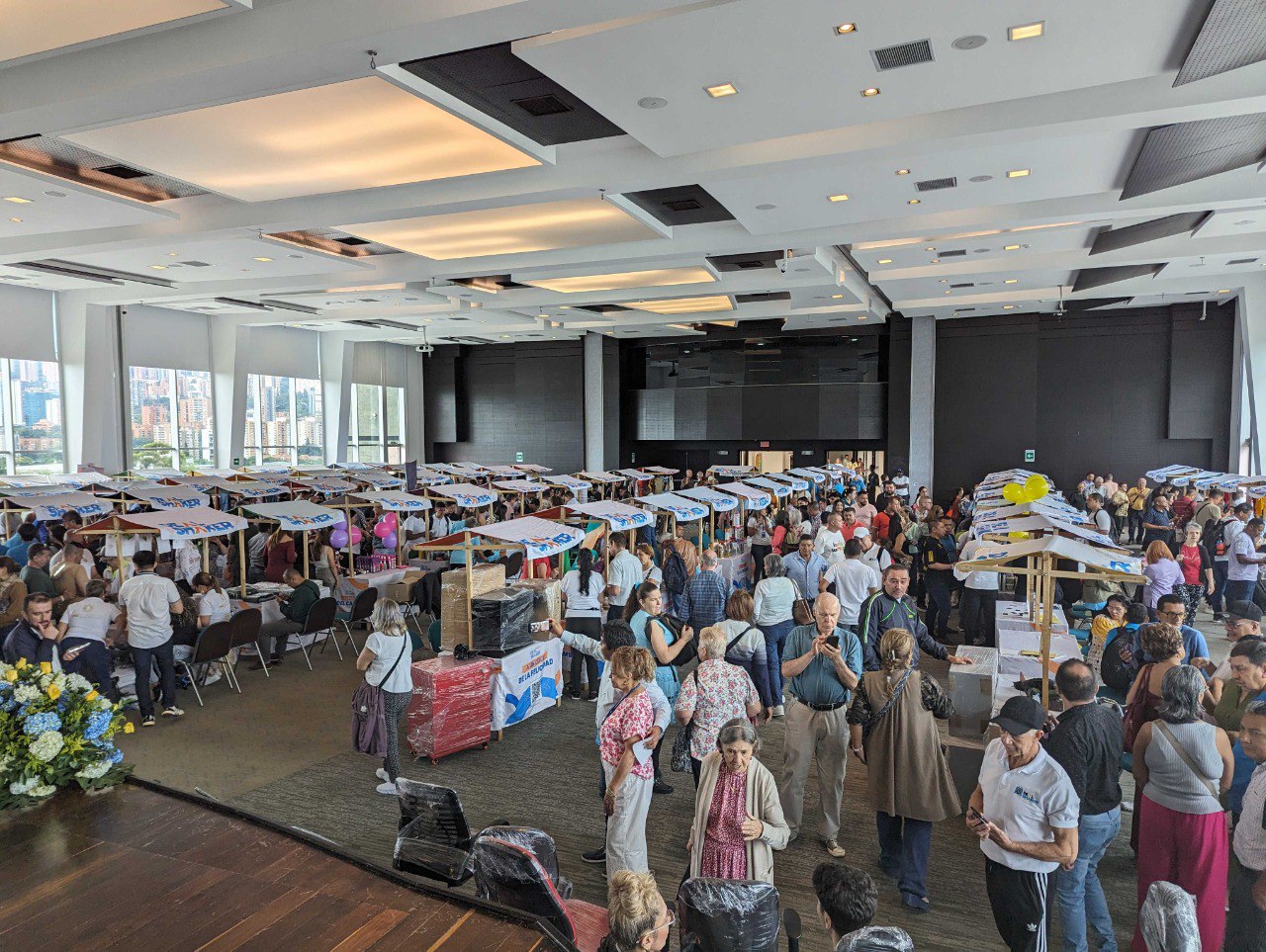 A CE é um evento indoor de grande escala onde um grande número de pessoas fica de pé e conversa em torno de uma mesa com vários itens em exibição em uma sala com grandes janelas e vista para a cidade.